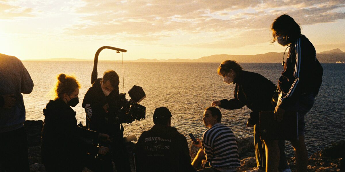 A film crew stands on the beach in front of a sunset.