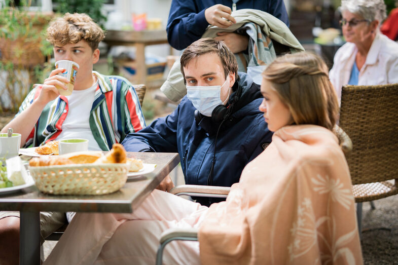 Two young people are sitting at a table in a café, with a person wearing a protective mask squatting between them.