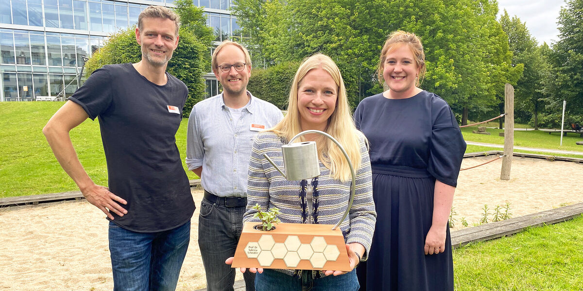 Vier Personen stehen auf dem Campus der FH Dortmund, eine Person hält einen Preis-Pokal in den Händen.