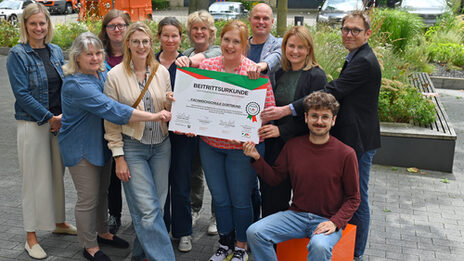 Personen stehen in einer Gruppe zusammen und halten eine große Urkunde von "Sicher im Dienst"