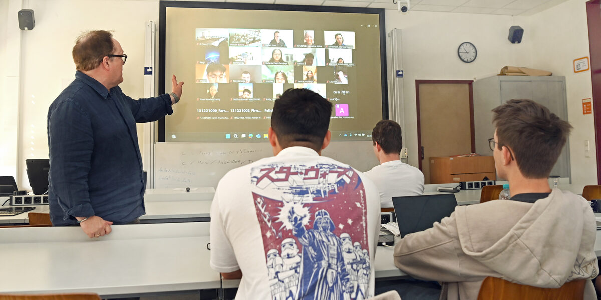 Menschen sitzen in einem Seminarraum und schauen auf eine Leinwand auf der weiter Personen zugeschaltet sind.