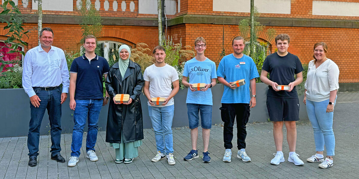 Eight people stand next to each other in front of a UAS building, some holding small boxes in front of them.