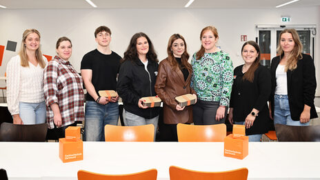 Acht Personen stehen in einem hellen, großen Raum hinter einem weißen Tisch und schauen freundlich in die Kamera.