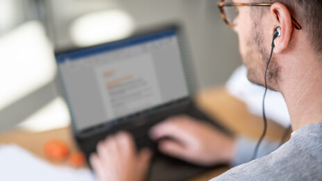 Foto eines Mannes am Laptop im Homeoffice mit Kopfhörern im Ohr. __ Man at laptop in home office with headphones in ear.