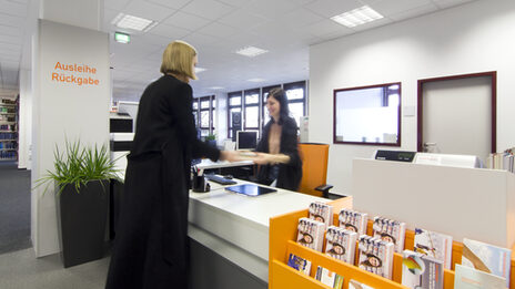 Foto einer Studierenden, die einer Mitarbeiterin der Bibliothek an der Ausleihtheke ein Buch übergibt__Photo of a female student handing a book to a female member of library staff at the service counter