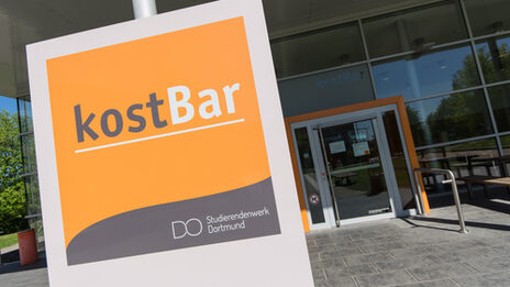 Photo of a sign with the logo of the kostBar canteen, in the background the entrance to the kostBar.