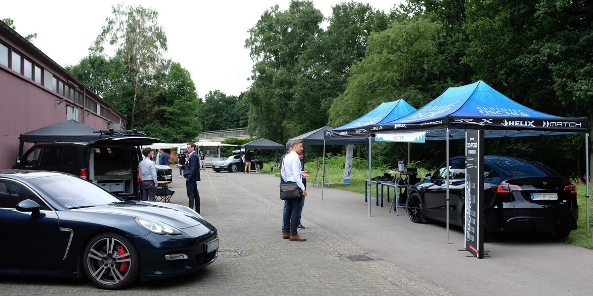Several exhibitor pavilions stand on a street next to a factory hall, with modern cars parked under some of them and a few people in between.