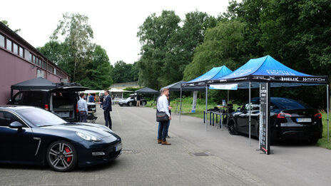 Mehrere Aussteller-Pavillons stehen an einer Straße neben einer Werkshalle, unter einigen von ihnen stehen moderne Autos, dazwischen einige Menschen.