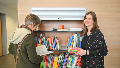 Two women are standing in front of a bookshelf. The woman on the left has her back to the camera and is reading a book. The woman on the right holds a stack of books in her hands and smiles at the camera.__ Two women are standing in front of a book shelf. The woman on the right has her back turned to the camera and is reading a book. The woman on the right is holding a pile of books and smiling at the camera.