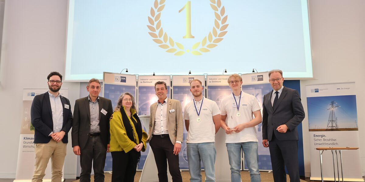 Seven people stand next to each other in front of the IHK's partition walls, with a golden emblem with the number 1 above them on a projector screen.