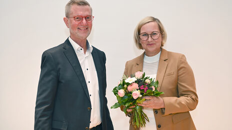 Eine männliche gelesene Person steht neben einer weiblich gelesener Person, die einen Blumenstrauß in der Hand hält.