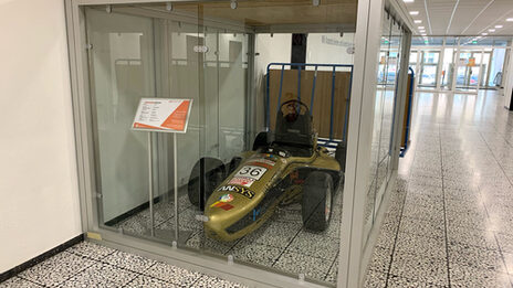 A racing car stands in a glass box next to a wall in a spacious hallway.