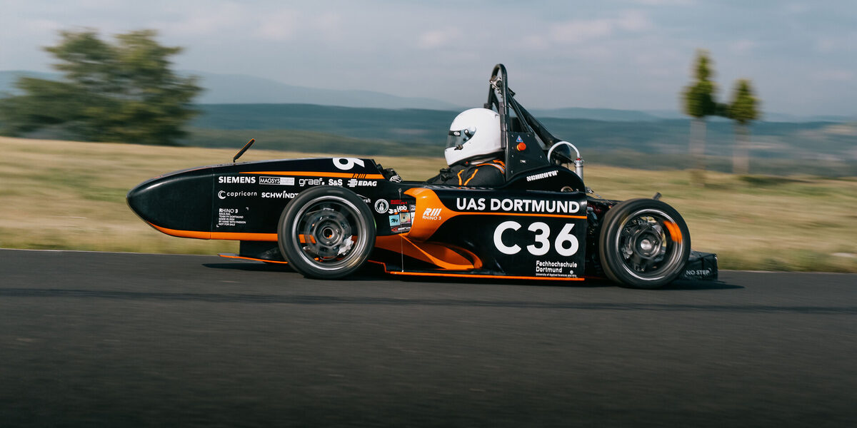 A person wearing a helmet is driving a racing car, the landscape can be seen blurred in the background.