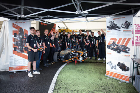 A group of people are standing under a large tent, with a racing car in front of them and two roll-ups next to them showing details of the car.