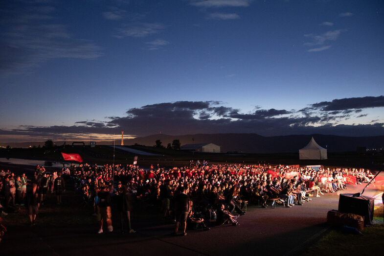 A large crowd is sitting and standing outside, some of the people are illuminated by spotlights, while a race track can be seen behind them in the twilight.