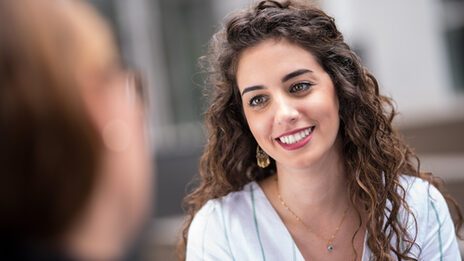 Closeup-Aufnahme einer jungen Frau mit dunklen lockigen Haaren, die eine Frau die ihr gegenüber sitzt anstrahlt.__Closeup of a young woman with dark curly hair smiling at a woman sitting across from her.