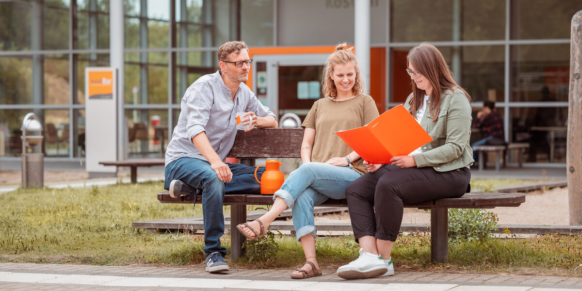 Foto von 3 Personen, die draußen vor der kostBar auf einer Holzbank sitzen und sich besprechen.