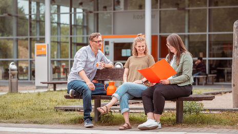 Photo of 3 people sitting on a wooden bench outside the kostBar and talking.