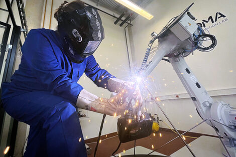 Links ein Mensch im blauen Arbeitsanzug mit Schweißhelm. Er schweißt die Enden mehrerer Stäbe zusammen. Rechts ein Roboterarm, der einen der Stäbe in Position hält. In der Mitte der Schweißpunkt mit Funkenregen.