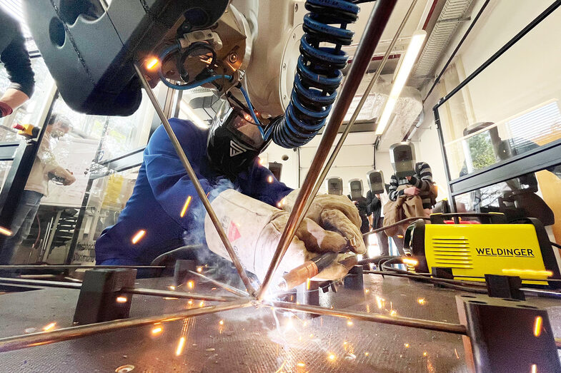 Im Vordergrund berühren sich die Enden mehrerer Metallstäbe. Ein Mensch hockt dahinter und schweißt sie zusammen. Von oben ragt ein Teil eines Roboterarms ins Bild, der einen der Stäbe hält. Im Hintergrund stehen mehrere Menschen mit Lichtschutzmasken und schauen zu.