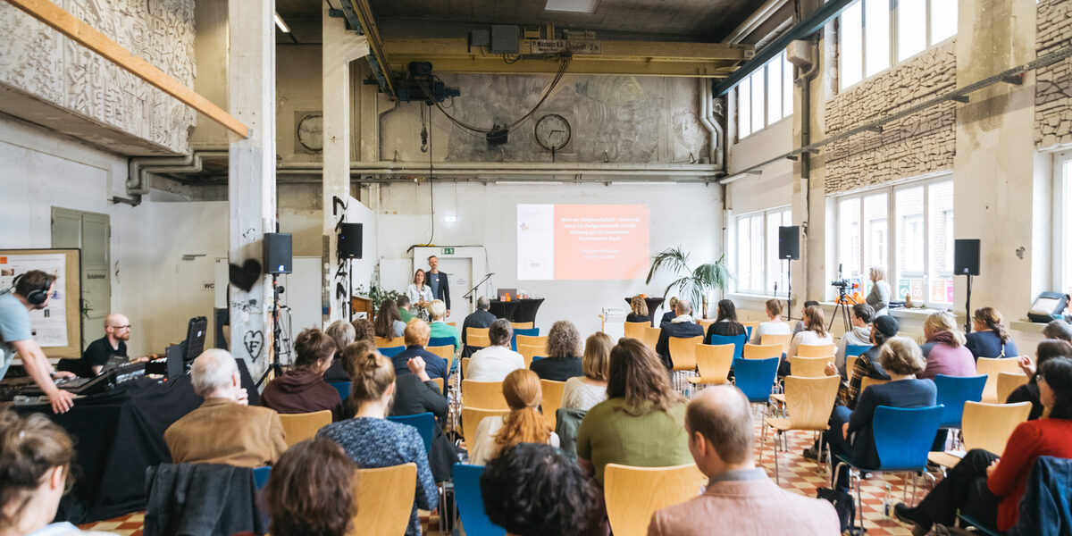 Blick in eine Halle, auf Stühlen sitzen Menschen (von hinten gesehen), vorne stehen zwei Personen dem Publikum zugewandt.