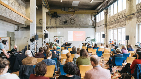 View into a hall, people are sitting on chairs (seen from behind), two people are standing in front facing the audience.