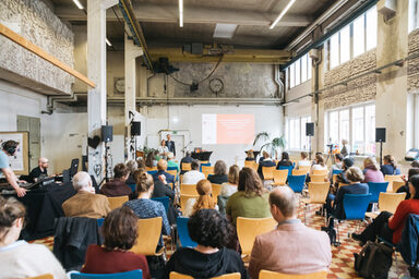 Blick in eine Halle, auf Stühlen sitzen Menschen (von hinten gesehen), vorne stehen zwei Personen dem Publikum zugewandt.