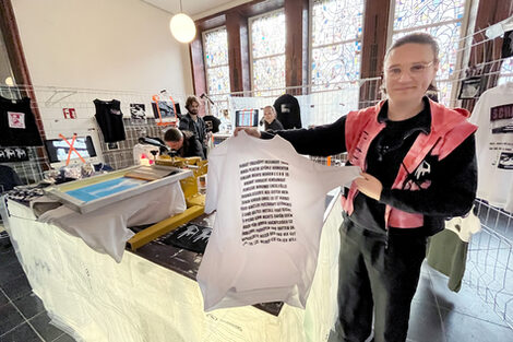 A man in black clothing and a red vest holds a T-shirt with words printed on it into the picture. He is standing at a stand with a screen printing machine and other items of clothing.