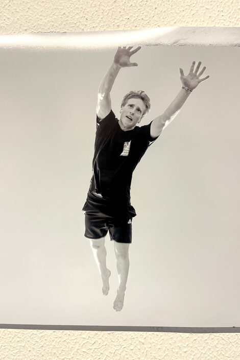 Black and white photo of a person in short, black clothing seen from below, performing a pike jump barefoot.