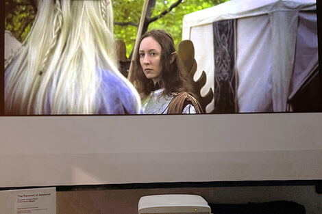 Movie scene of two elves looking into each other's eyes on a window-sized screen in front of a wall, with a low stool with a projector in front of it.