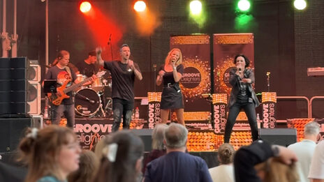 Frontalblick über die Köpfe einiger Menschen hinweg auf eine Bühne, auf der ein Bassist, ein Schlagzeuger und drei Sänger*innen in Aktion zu sehen sind.