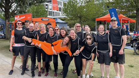 15 Personen in schwarzen Trikots tragen eine weitere Person in Orange auf Händen, stehen nebeneinander und schauen in die Kamera. Im Hintergrund stehen ein Auto und zwei Zelte, beide orange, auf einer Wiese.