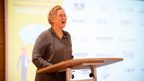 A person with a head microphone stands at a lectern and speaks. A projection can be seen in the background.