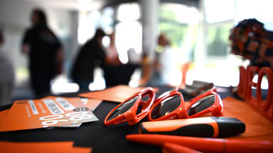 Orangefarbene Sonnenbrillen, Stifte und Postkarten liegen auf einer schwarzen Oberfläche, im Hintergrund sind unscharf mehrere Personen zu erkennen.