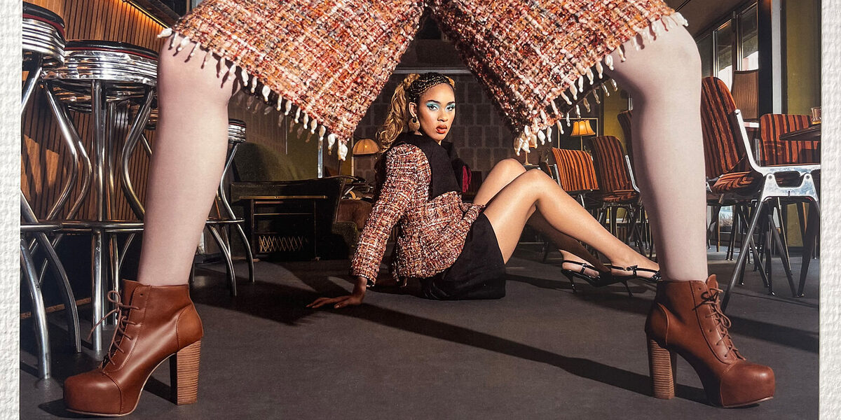 Durch die Beine einer Person im Vordergrund ist eine dahinter auf dem Boden sitzende weitere Person zu sehen. Beide Personen sind schick gekleidet.