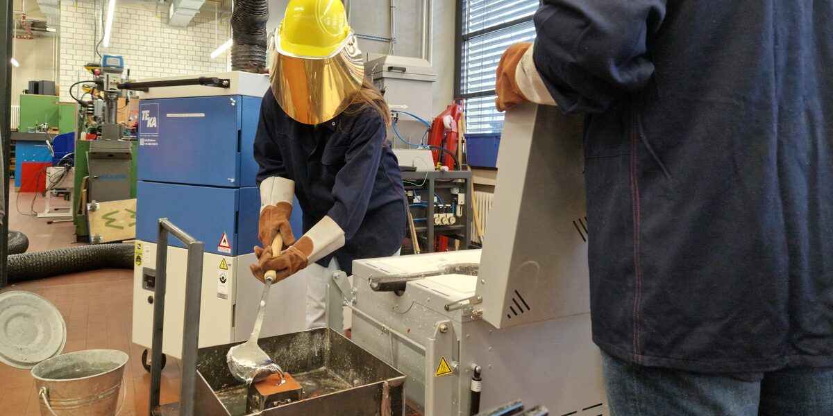 A person fills a silvery substance into a container with a ladle. The person wears gloves and a protective helmet with a visor that covers the entire face.