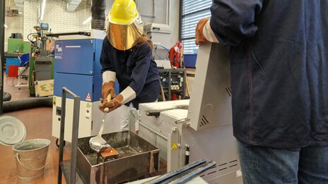 A person fills a silvery substance into a container with a ladle. The person wears gloves and a protective helmet with a visor that covers the entire face.