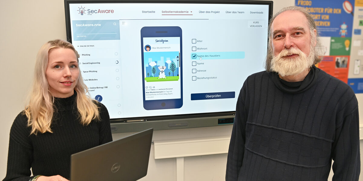 Two people are standing in front of a large screen showing the digital learning platform "SecAware".nrw.