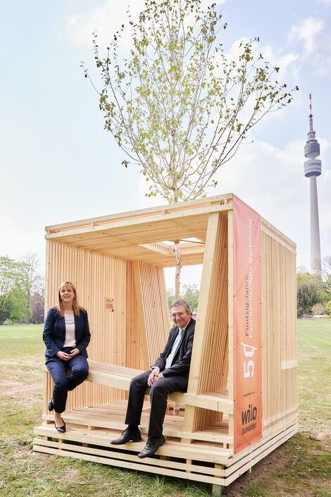 Zwei Menschen sitzen in einem großen HolzwürFHel. Im Hintergrund ist der Fernsehturm des Westfalenparks zu sehen.