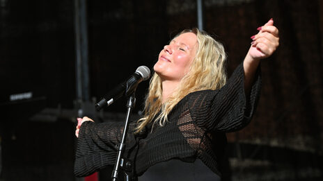 A person with long blond hair and outstretched arms stands in front of a microphone with closed eyes and an inward smile.