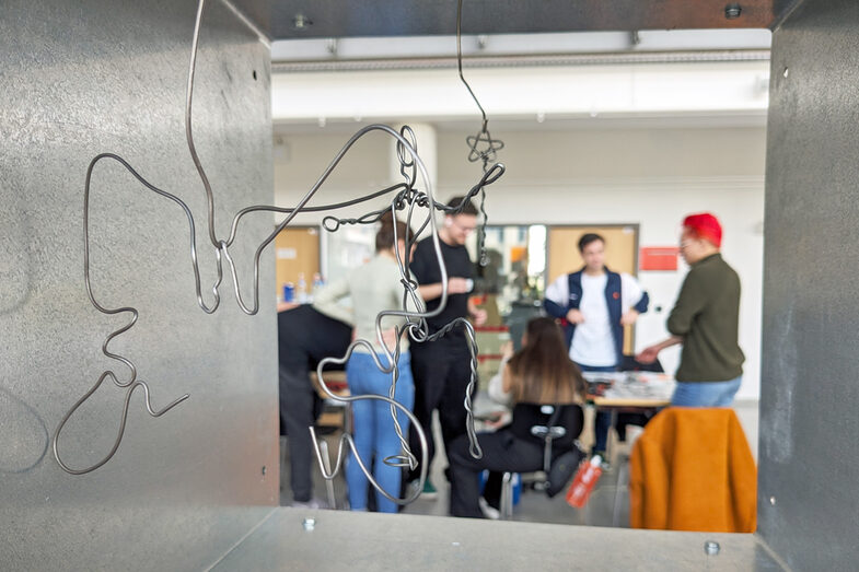 A wire construction hangs in a metal frame, in the background people are working in a team.