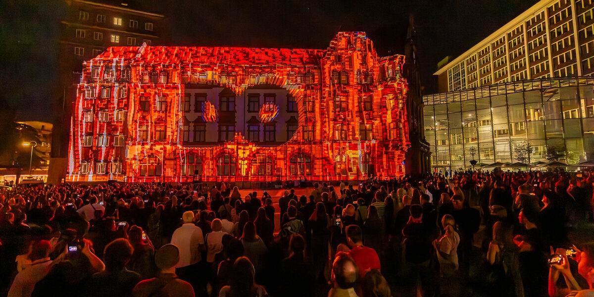 Ein Gebäude ist komplett orange angestrahlt, in der Mitte sind die Buchstaben DO zu lesen. Davor stehen unzählige Menschen im Dunklen.