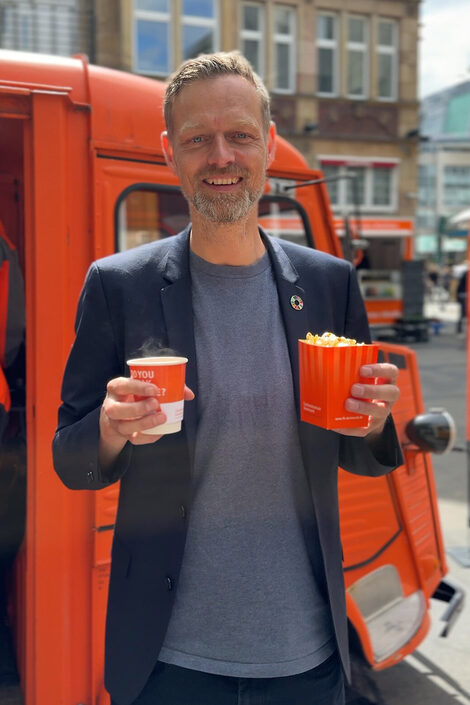 Eine Person steht mit einem Pappbecher und einer Packung Popcorn vor einem Oldtimer.