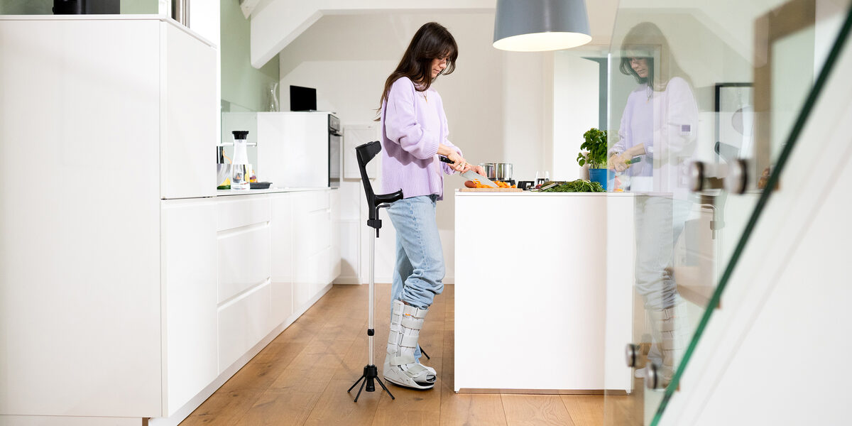 A person read as female is standing in a kitchen. She has her right foot in a splint. Next to her is the walking aid with the STEETS module.