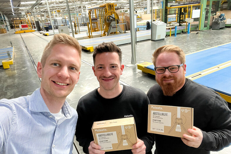 Three people are standing in a production hall, two are holding a cardboard box in their hands.