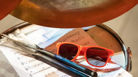 Photo of a drum kit. On it are a sheet of music, a pair of sunglasses and two drumsticks.