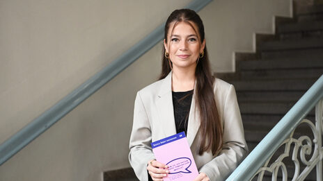 Ein Mensch im hellen Blazer steht auf einer Treppe, schaut in die Kamera und hält ein Buch in den Händen.