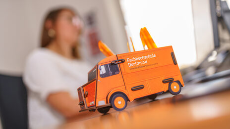 Photo of a pen box in the form of a Citroen HY panel van on a desk, in the background a woman is working on the computer. __ In the foreground on the desk is a pen box in the form of a Citroen HY panel van, in the background a woman is working on the computer.