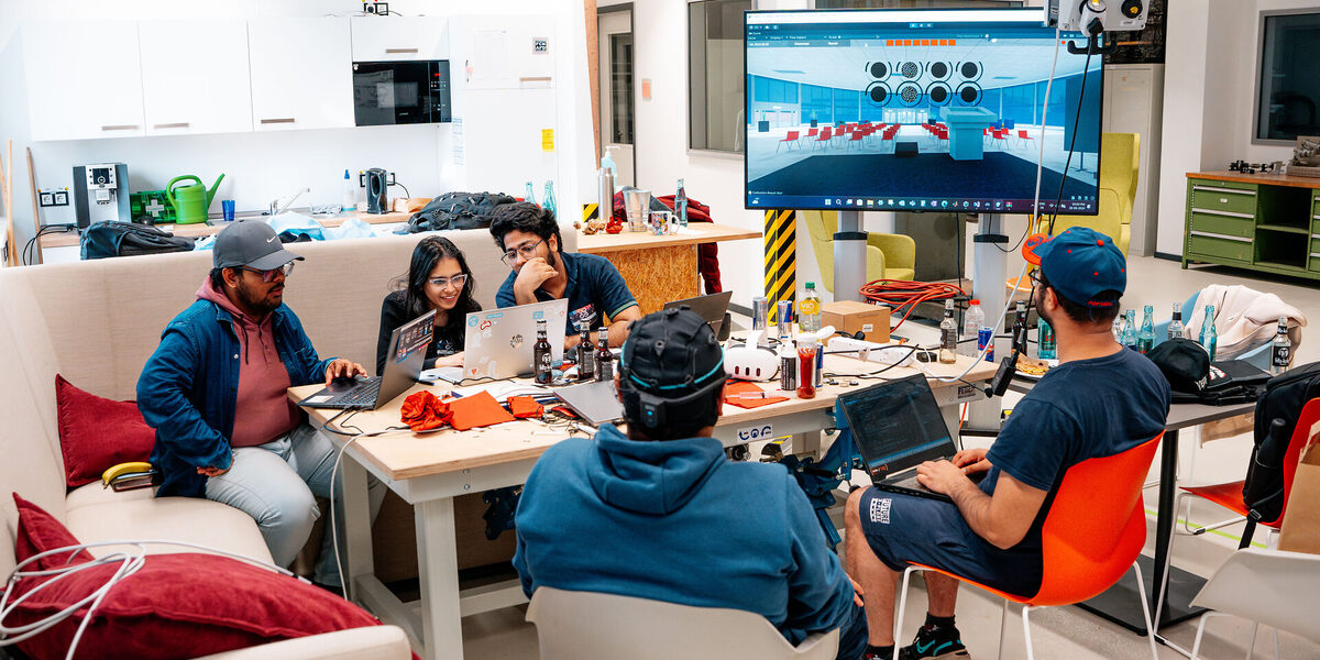 Five people sit in a room at a table with laptops and technical equipment, next to a large screen showing a virtual room with circle symbols.