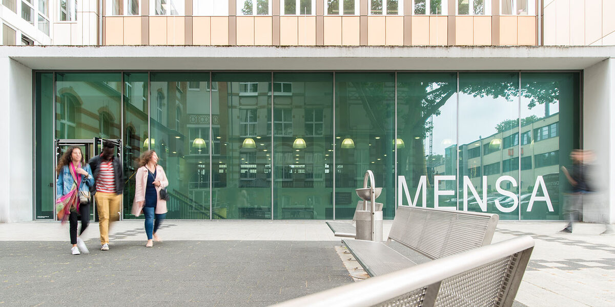 Drei Studierende kommen aus der neuen Mensa an der Sonnenstraße. Groß im Bild ist die Glasfassade der Mensa.